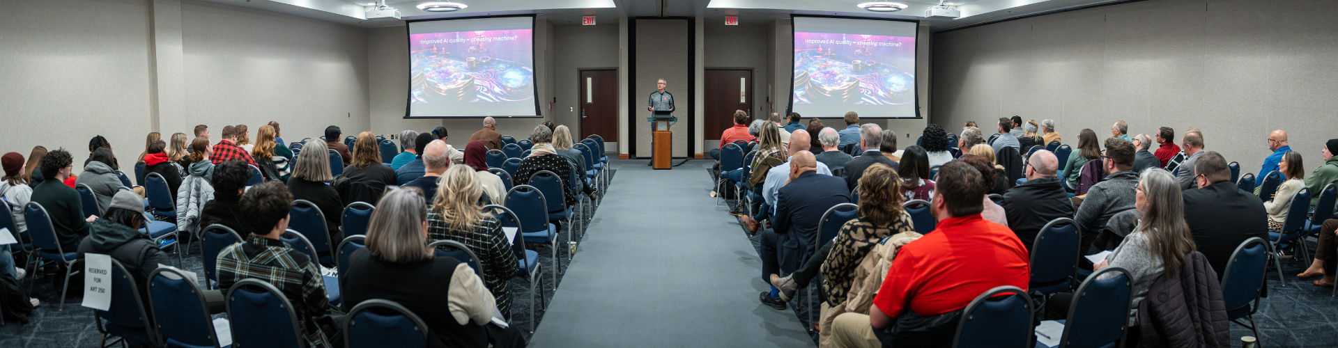 crowd seated in theater style rows of chairs facing a presenter and two projector screens